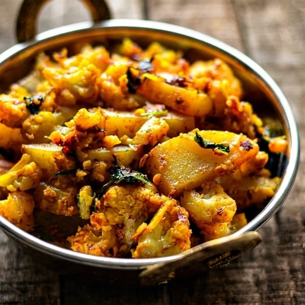 A metal bowl filled with golden-brown spiced cauliflower and potato curry on a rustic wooden table.