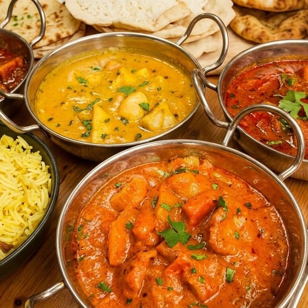 Five bowls of colorful Indian curries and yellow rice, with naan bread and cutlery on a wooden table.